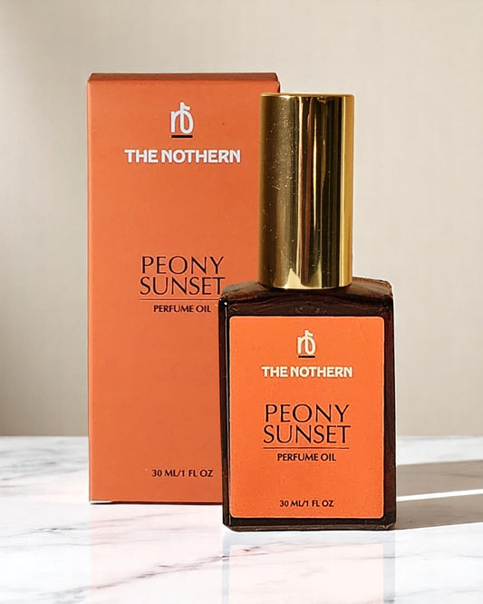 A bottle of Peony Sunset Perfume Oil by The Northern, with a gold cap over pump applicator, next to its orange packaging box.