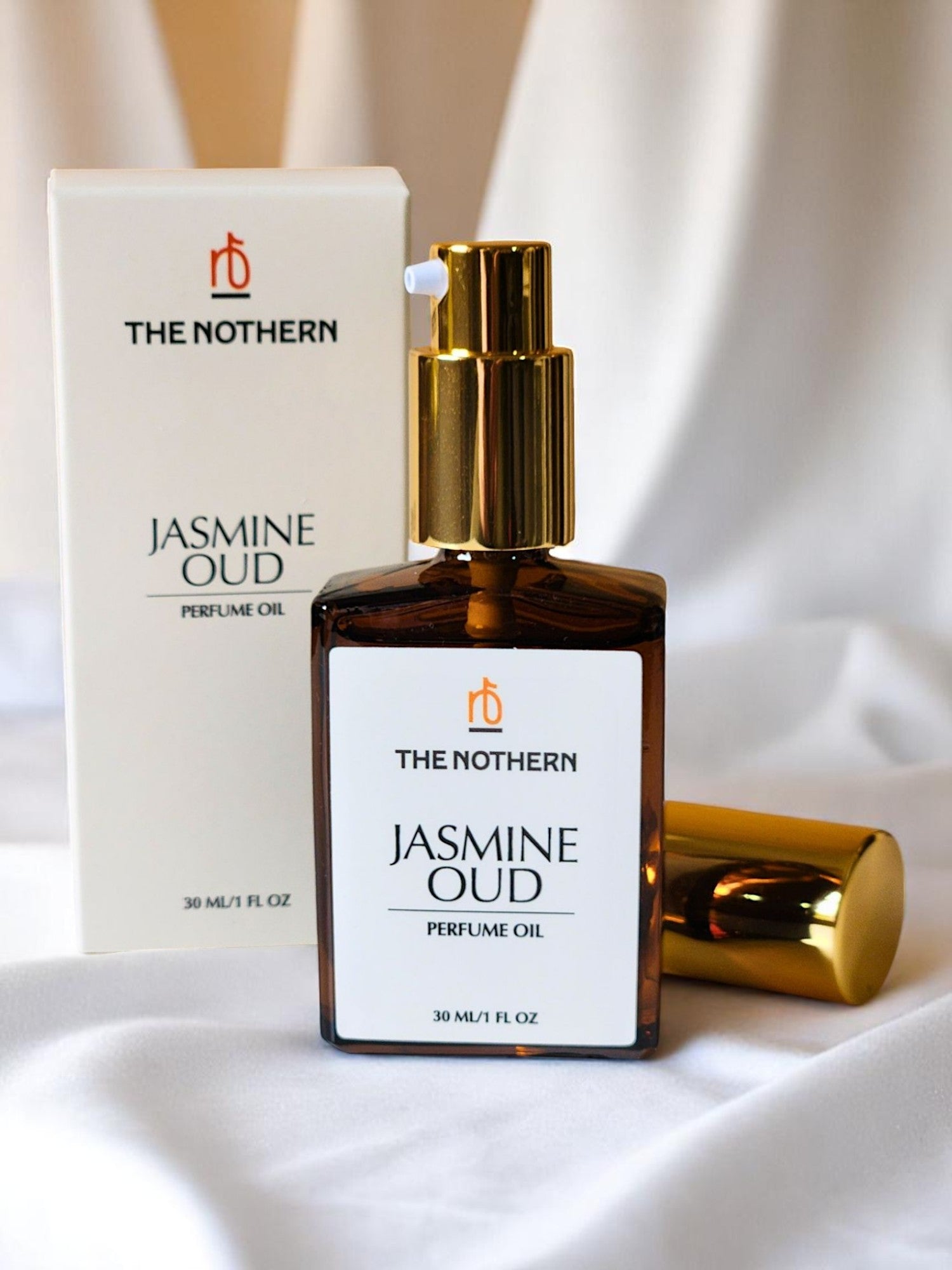 The Nothern Jasmine Oud Perfume Oil bottle with pump applicator next to top cover laying flat & packaging on draped white fabric background